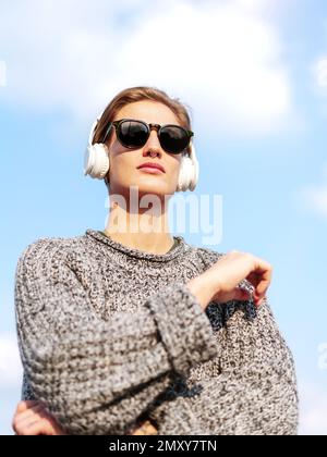 Selbstbewusste junge Frau mit Sonnenbrille und Kopfhörern in lässiger Kleidung, die gegen den blauen Himmel posiert. Stockfoto