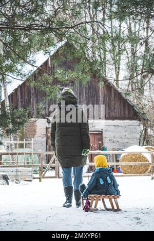 Mutter oder Mädchen oder Frau, die im Winter auf dem Land spazieren geht, mit Kind im Schlitten und weißer Winterlandschaft, Holzhaus, Farm, Heuballen Stockfoto