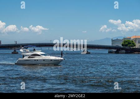 Ein weißes Schnellboot, das auf dem Wasser der Guanabara-Bucht segelt, nahe dem Sao Bento Pier mit der Arnaldo Luz-Brücke auf der Rückseite unter klarem Himmel im Sommer am Nachmittag. Stockfoto