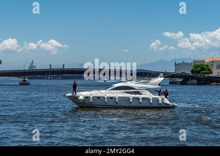 Ein weißes Schnellboot, das auf den Gewässern der Guanabara Bay segelt, nahe Sao Bento Pier mit Arnaldo Luz Brücke hinten unter dem sonnigen blauen Himmel. Stockfoto