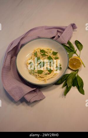 Cremige Nudelgerichte mit Basilikum-Dressing. Auf weißem Marmorhintergrund mit lila Geschirrtüchern, Zitrone und frischem Basilikum. Von oben genommen. Stockfoto