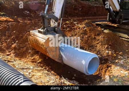 Zur Vorbereitung der unterirdischen Montage von Betonabwasserrohren unter dem Boden verwendet der Arbeiter einen Traktor, um diese Rohre anzuheben Stockfoto