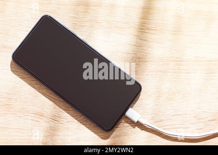 Zenithale Aufnahme eines Mobiltelefons, das mit einem weißen usb-Kabel über einem Holztisch aufgeladen wird. Stockfoto