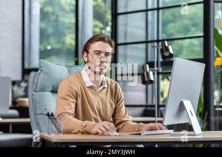 Porträt eines jungen Mannes, Psychologen, Psychotherapeuten, der im Büro sitzt, am Computer arbeitet, online konsultiert, per Videoanruf mit Patienten spricht. Er schaut ernsthaft in die Kamera. Stockfoto