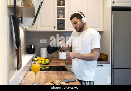 Mann mit kabellosen Kopfhörern und weißem T-Shirt steht in der Küche, gießt Kaffee in die Tasse und hört Musik. Stockfoto
