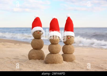 Schneemänner aus Sand mit Weihnachtsmannmützen am Strand nahe dem Meer. Weihnachtsferien Stockfoto