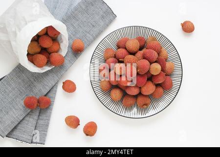 Lychee-Früchte auf asiatischem Teller auf weißem Hintergrund mit ganzen Stücken, Blick von der Tischplatte. Frisches Litchi-Obst in Papiertüte, Foto direkt von oben. Stockfoto