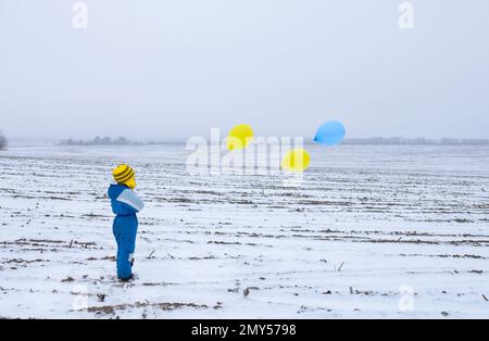 Nicht wiedererkennbarer einsamer Junge, der auf einem verschneiten Feld steht und gelbe blaue Ballons hält. Konzept der Aufmerksamkeit auf Krieg, militärischen Terror in der Ukraine. Gestohlen Stockfoto