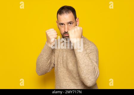Ein hübscher bärtiger Mann mit Rollkragenpullover auf gelbem Hintergrund. Faust zum Kampf schlagen, aggressiver und wütender Angriff, Bedrohung und Gewalt. Stockfoto