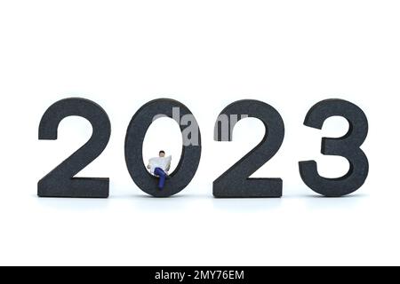 2023 Business Neujahrskonzept. Nahaufnahme von Miniaturfiguren aus dem Geschäftsleben, die eine Zeitung auf einer Holznummer auf weißem Hintergrund lesen. Stockfoto