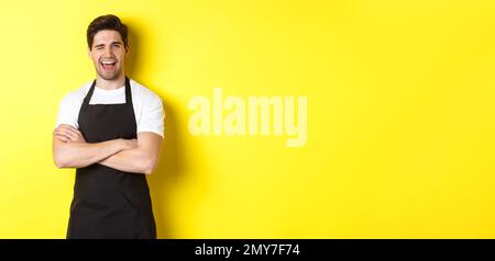 Ein hübscher Barista in schwarzer Schürze winkend zu dir, trägt schwarze Schürzenuniform und steht auf gelbem Hintergrund Stockfoto