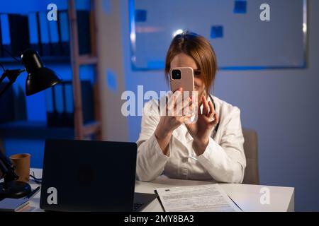 Eine Ärztin, die sich ein Smartphone ansieht und nachts am Büroschreibtisch mit einem Laptop arbeitet, Telefonkonferenzen, e-Health-Service. Health App ein Stockfoto