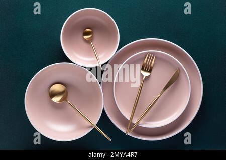 Geschirr und goldenes Besteck auf dunkelgrünem Hintergrund, Blick vom Tisch, oben. Flach liegendes Design. Leere Keramikteller, Utensilien zum Servieren. Registerkarte „Trend“ Stockfoto