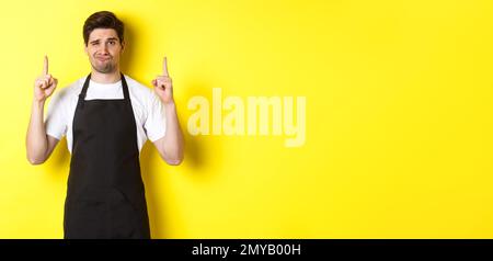 Unzufriedener und skeptischer Kellner mit schwarzer Schürze, der die Finger nach oben zeigt, drückt Abneigung und Gleichgültigkeit aus und steht auf gelbem Hintergrund Stockfoto
