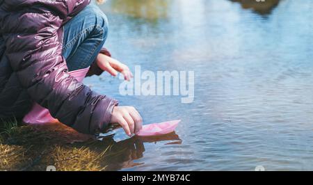 Mädchen in lila Jacke, die ein Papierboot startet. Farbenfrohes pinkfarbenes Schiff in den Händen. Kinder spielen, haben Spaß im Freien in der Frühlingsschneepfütze auf der Straße. Krieg Stockfoto