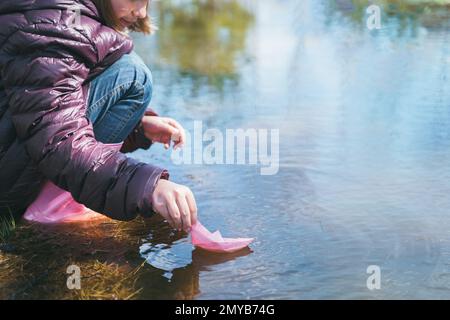 Mädchen in lila Jacke, die ein Papierboot startet. Farbenfrohes pinkfarbenes Schiff in den Händen. Kinder spielen, haben Spaß im Freien in der Frühlingsschneepfütze auf der Straße. Krieg Stockfoto