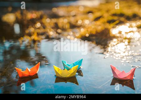 Mehrfarbige Papierboote. Farbenfrohe rosafarbene, blau-orange Schiffe in einer großen Schneepfütze im Frühling auf der Winterstraße. Warmes, nasses Regenwetter, altes Gras. Hallo Stockfoto