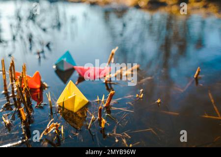 Mehrfarbige Papierboote. Farbenfrohe rosafarbene, blau-orange Schiffe in einer großen Schneepfütze im Frühling auf der Winterstraße. Warmes, nasses Regenwetter, altes Gras. Hallo Stockfoto