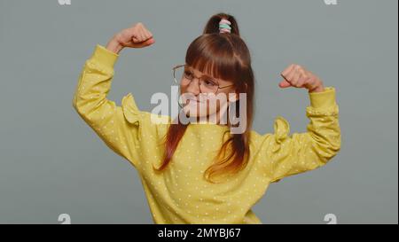 Ich bin stark und gesund. Ein junges Mädchen mit Brille, das Bizeps zeigt und selbstbewusst aussieht. Es fühlt Kraft, um für Rechte zu kämpfen, Energie, um erfolgreich zu gewinnen. Ein Kind mit grauem Hintergrund Stockfoto