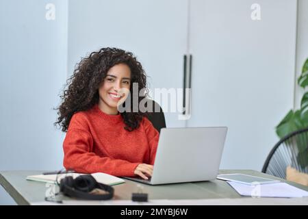 Junge lächelnde lateinische Geschäftsfrau, die mit einem Laptop am Schreibtisch sitzt. Stockfoto