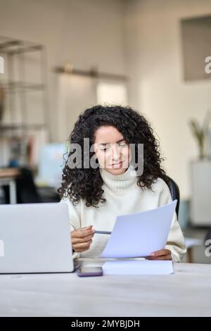 Junge lateinische Geschäftsfrau mit Dokumenten, die an einem Laptop arbeiten, vertikal. Stockfoto