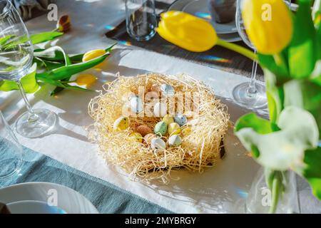 Schöne Ostertische mit festlichem Dekor im Innenbereich. Serviette in Form von Kaninchenohren, bunte Eier im Nest, frische gelbe Tulpen in Vase. Sonnenlicht Stockfoto