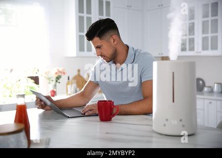 Mann mit Laptop in der Küche mit modernem Luftbefeuchter Stockfoto