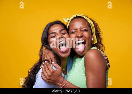 Zwei Diversität ziemlich positive Mädchen, die dir die Zunge ausstechen, isoliert auf gelbem Hintergrund umarmt. Stockfoto