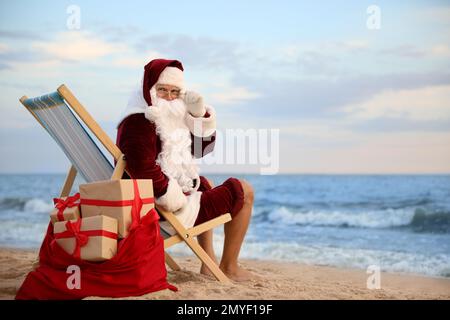 Weihnachtsmann mit Geschenktüte zum Entspannen im Sessel am Strand, Platz für Text. Weihnachtsferien Stockfoto