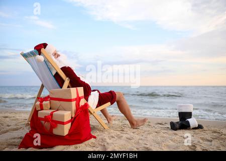 Weihnachtsmann mit Geschenktüte zum Entspannen im Sessel am Strand, Platz für Text. Weihnachtsferien Stockfoto