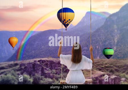 Traumwelt. Eine junge Frau, die über Berge schwingt, Heißluftballons am Sonnenuntergang im Hintergrund Stockfoto