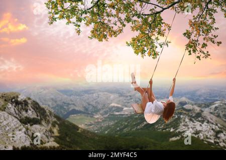 Traumwelt. Eine junge Frau, die unter dem Sonnenuntergang über den Bergen schwingt Stockfoto