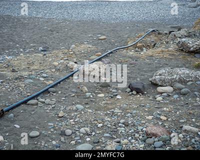 Kabel auf schwarzem Sand. Das Kabel ragt aus dem Boden heraus. Elektrische Kommunikation am Strand. Industrie. Unfertige Arbeit Stockfoto