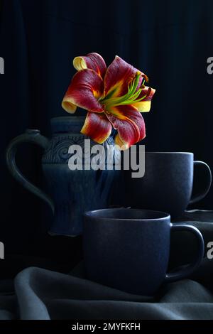 Stillleben mit Bechern und Gläsern, mit einer hellgelb-orangefarbenen Lilly auf dunklem Hintergrund. Stockfoto