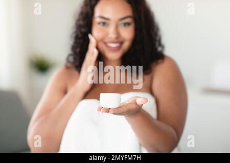 Glückliche schwarze, übergewichtige Frau, die ein Glas mit organischer Hautcreme hält und nach dem Duschen Feuchtigkeitscreme auf das Gesicht aufträgt Stockfoto