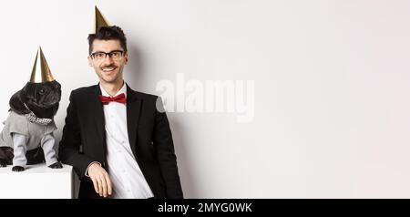 Bild von schönen jungen Mann Geburtstag feiern mit niedlichen schwarzen Mops in Party-Kostüm und Kegel auf dem Kopf, stehen über weißem Hintergrund Stockfoto