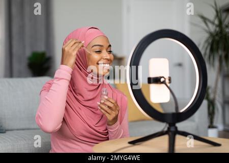 Fröhliche, schwarze, muslimische Schönheitsbloggerin, die ein Video auf einem Smartphone macht, auf einer Ringlampe, die ein neues Gesichtsserum zeigt Stockfoto