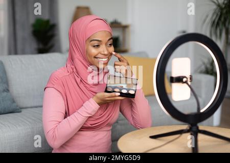 Eine glückliche schwarze moslem-Make-up-Künstlerin sitzt vor einem Handy, das auf einer Ringlampe sitzt, und strömt von zu Hause Stockfoto