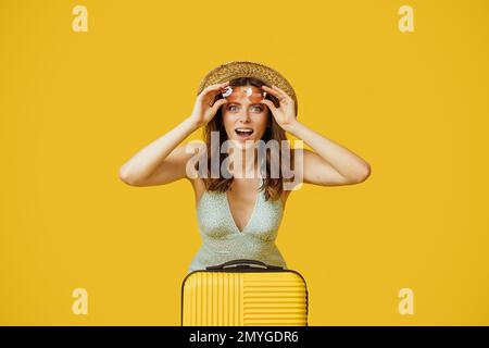 Erstaunte Frau in Kleid und Strohhut mit Sonnenbrille, die mit offenem Mund in die Kamera schaut, mit Koffer sitzt Stockfoto
