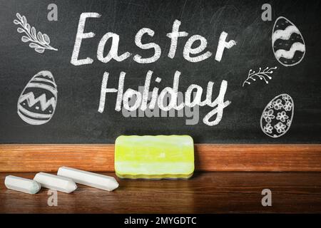 Text Osterfeiertag und Zeichnungen auf schwarzer Tafel neben Holztisch. Schulferien Stockfoto