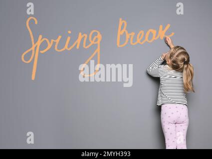 Ein kleines Mädchen schreibt einen Satz Spring Break mit Kreide an der grauen Wand. Schulferien Stockfoto