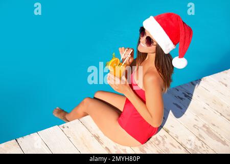 Junge Frau mit Weihnachtsmann-Hut und erfrischendem Getränk am Pool. Weihnachtsferien Stockfoto