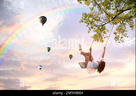 Traumwelt. Junge Frau schaukelt, Heißluftballons am Sonnenuntergang im Hintergrund Stockfoto