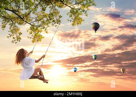 Traumwelt. Junge Frau schaukelt, Heißluftballons am Sonnenuntergang im Hintergrund Stockfoto