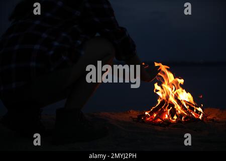 Eine Frau, die nachts am Strand neben brennendem Feuerholz die Hände wärmt, Nahaufnahme Stockfoto