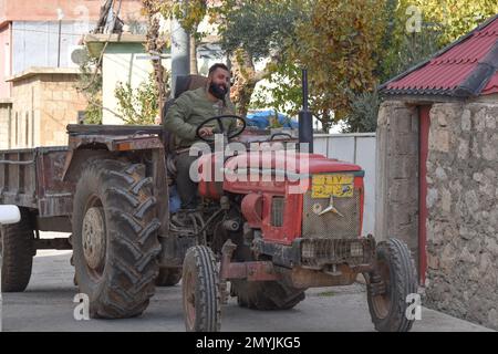 Ein lächelnder Mann, ein Handy in der Hand, führt seinen alten, roten Traktor, der einen Anhänger zieht, durch die Straßen eines Dorfes im Nordirak. Stockfoto