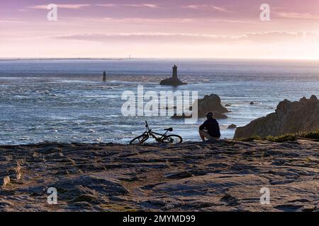 Radfahrer sitzt an der felsigen Küste, macht eine Pause, Pointe du Raz, Phare de la Vieille, Silhouette gegen den Abendhimmel, Cap Sizun, Département Finistère, Stockfoto