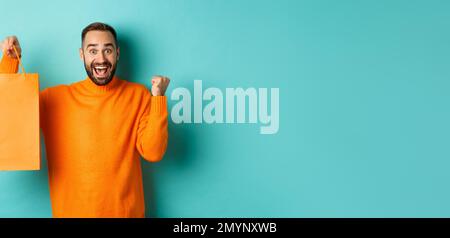 Glücklicher Mann hält orange Einkaufstasche und jubeln, bekam Rabatt und feiern, stehen über türkisfarbenem Hintergrund Stockfoto