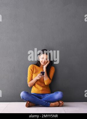 Ich überprüfe ihren Posteingang nach neuen Nachrichten. Eine attraktive junge Frau, die mit ihrem Handy auf dem Boden sitzt. Stockfoto