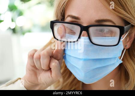 Frau, die neblige Brille abwischte, verursacht durch das Tragen einer medizinischen Maske in Innenräumen, Nahaufnahme Stockfoto